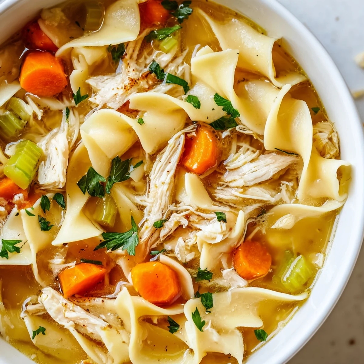 A close-up of Chicken Noodle Soup with Homemade Egg Noodles reveals golden broth, fresh parsley garnish, and rustic crusty bread on the side.