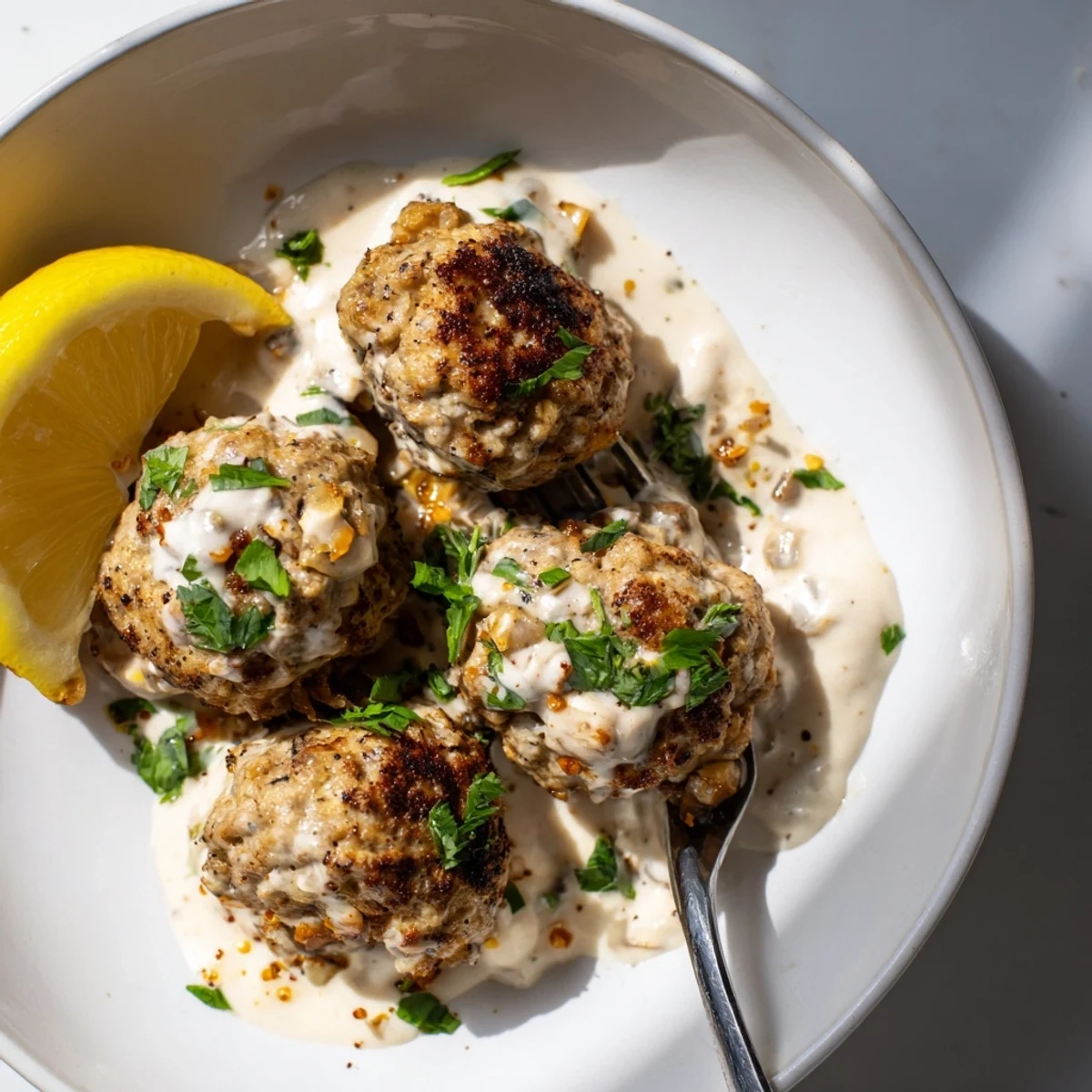 Juicy chicken kofta meatballs glisten with spices beside creamy garlic yogurt sauce in a white bowl.
