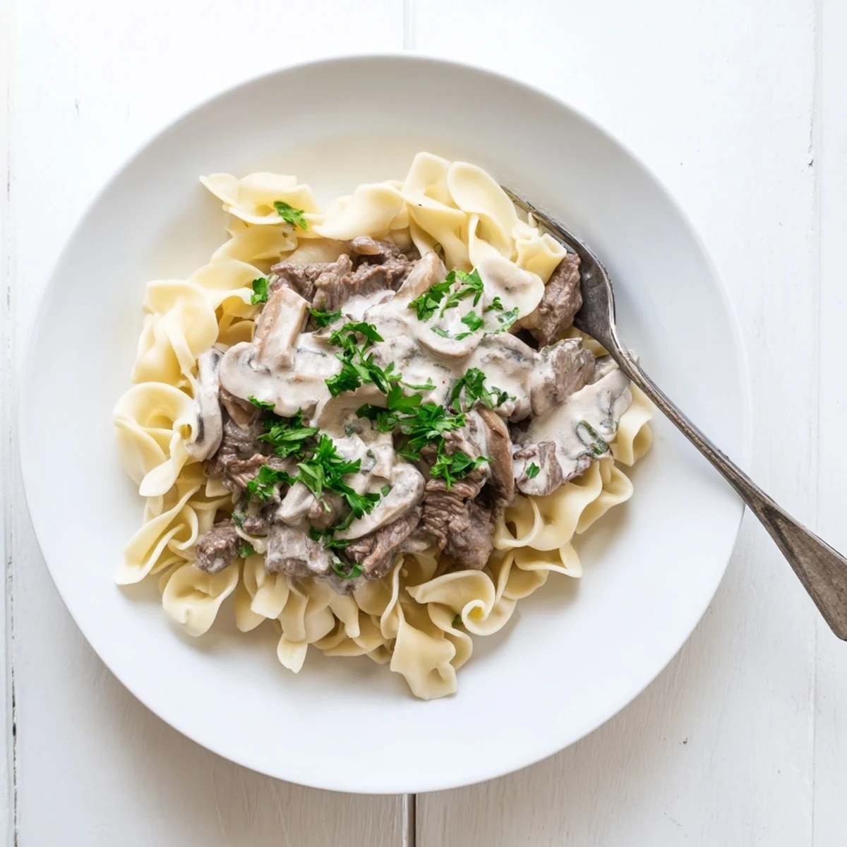 Homemade beef and mushroom stroganoff plated over egg noodles with chopped parsley sprinkled on top