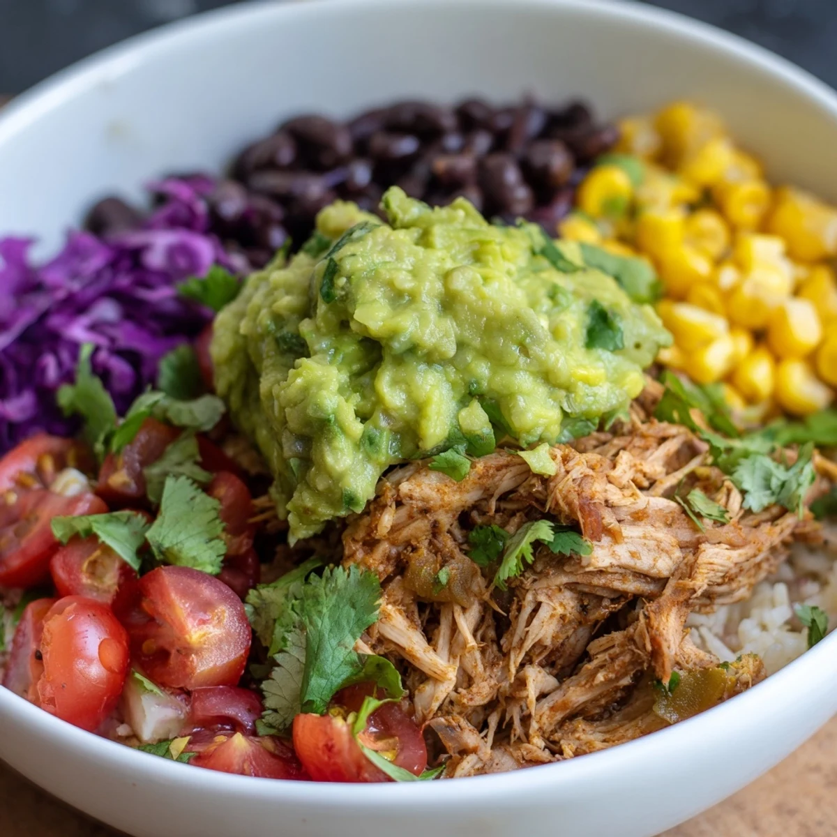 Healthy pulled chicken guacamole bowl featuring seasoned meat, ripe avocado mash, black beans, corn, and crisp red cabbage